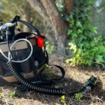 A large tool bag filled with tree healthcare tools, including a tree injection system, is lying on the ground next to some trees.