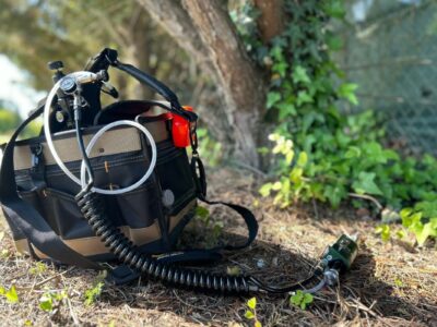 A large tool bag filled with tree healthcare tools, including a tree injection system, is lying on the ground next to some trees.